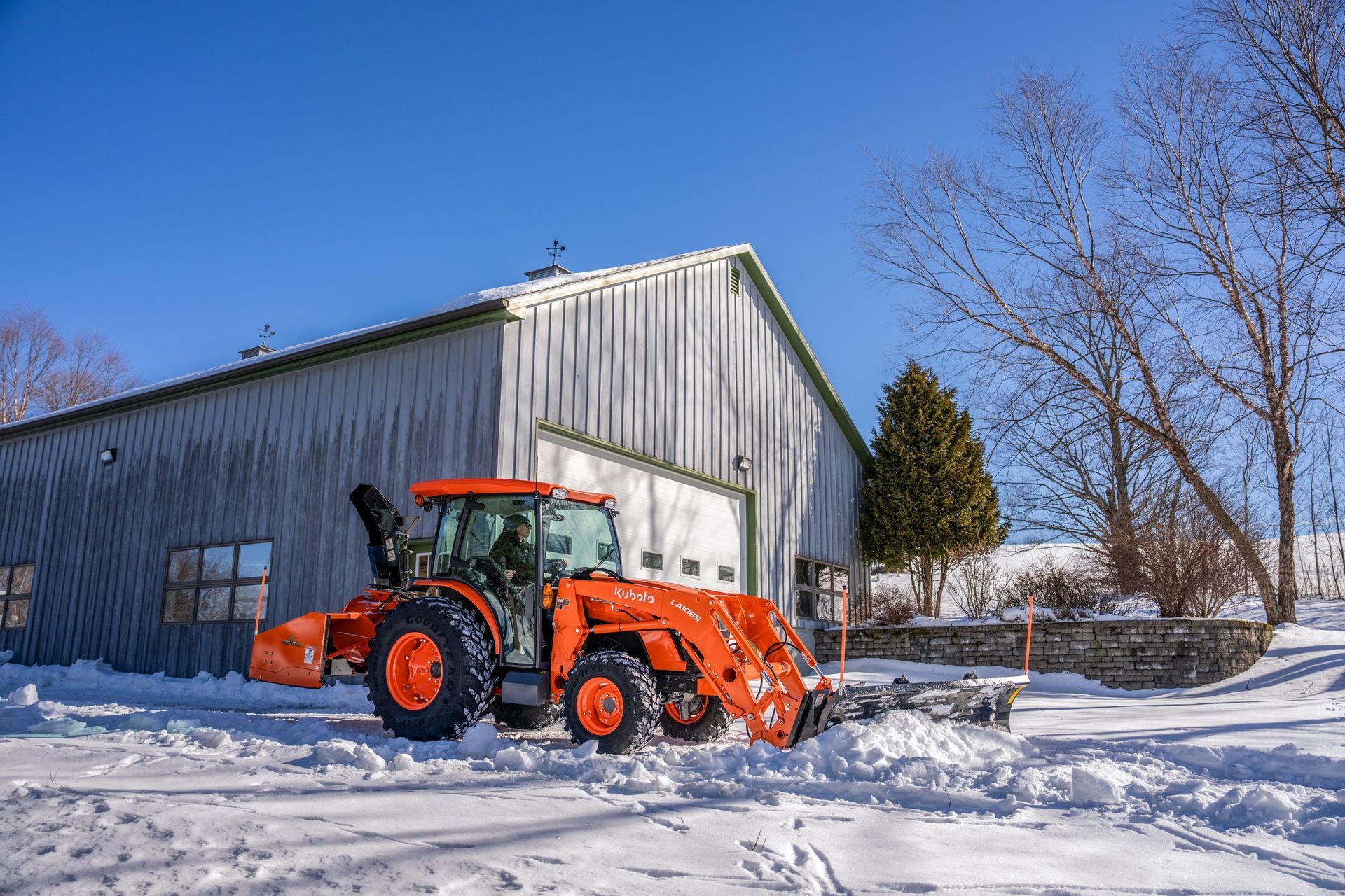 Orange tractor plowing snow in front of a metal-sided building on a sunny winter day.