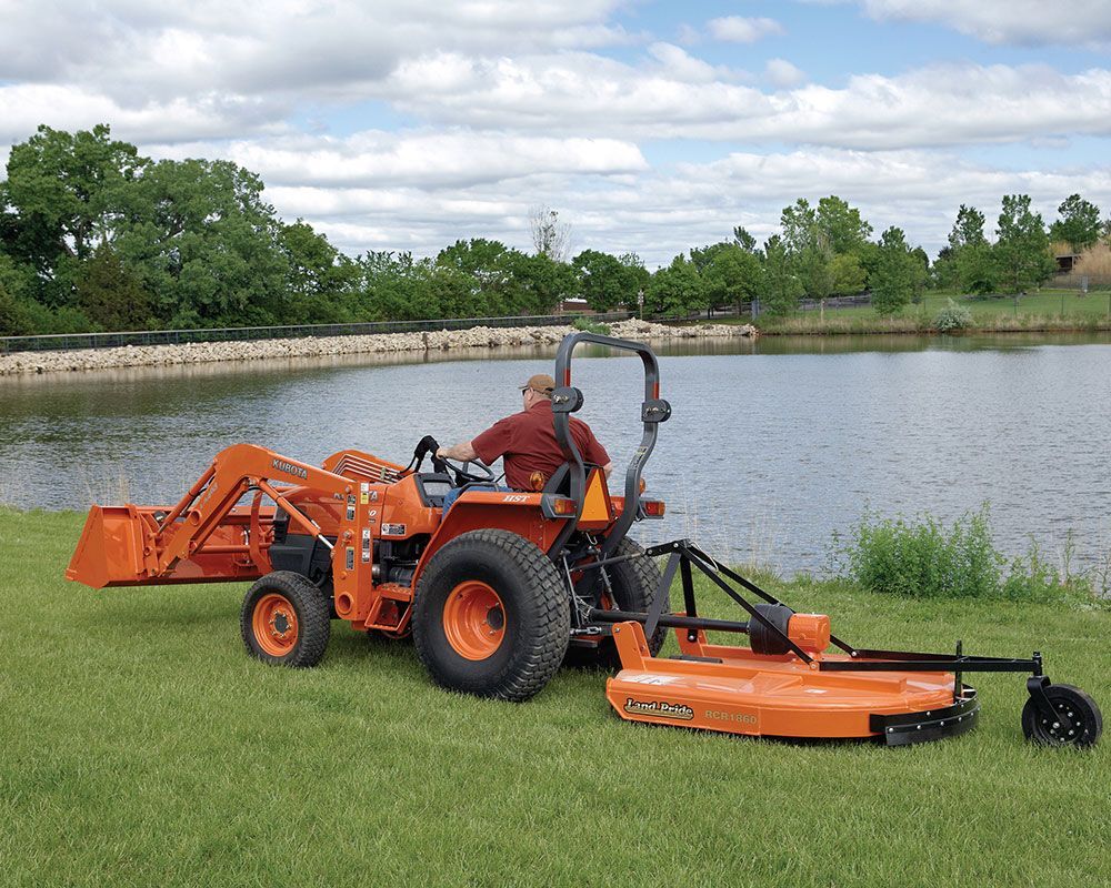 Orange tractor with a front loader and mower near a pond. A person operates the tractor on a grassy bank.