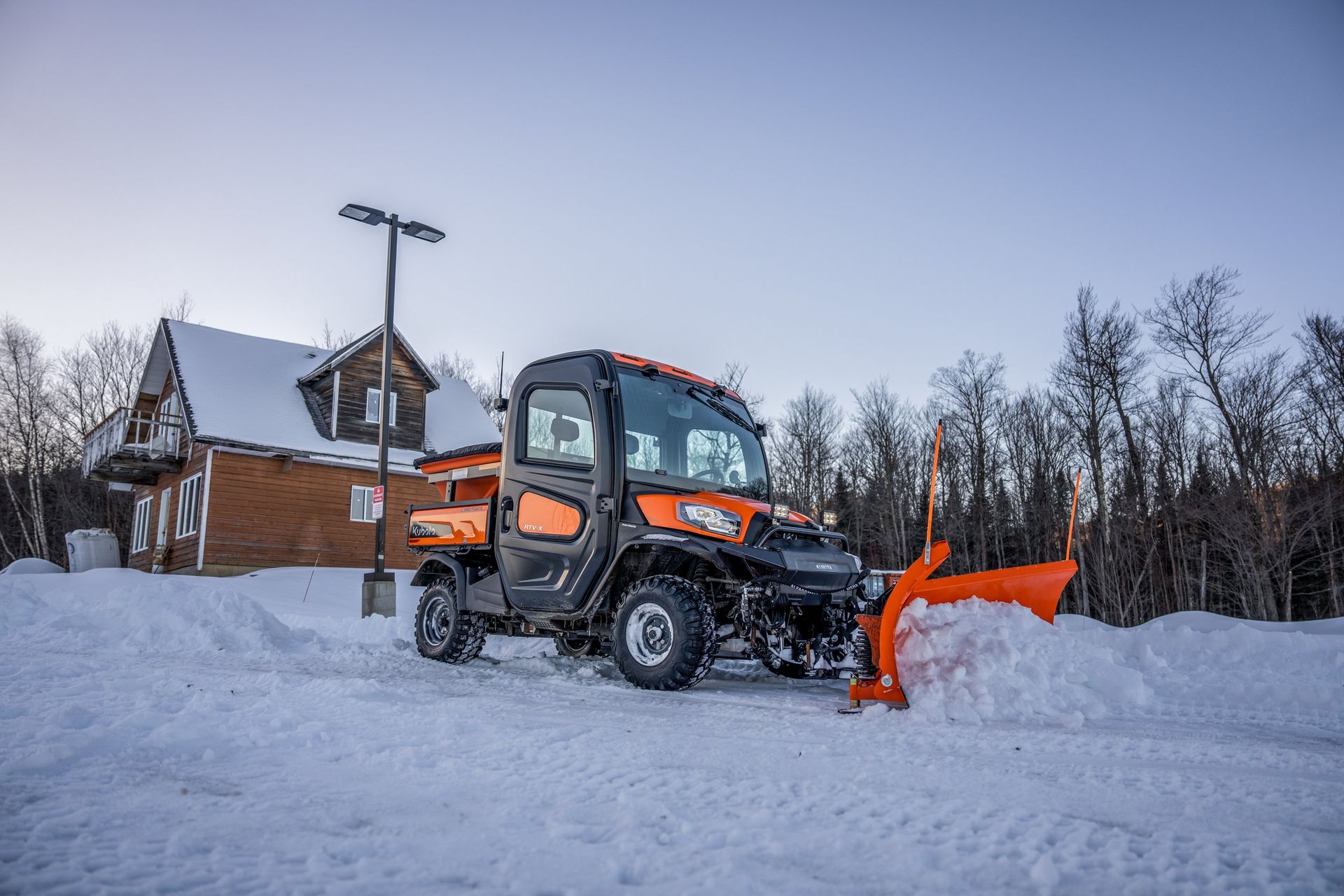 Orange utility vehicle with snowplow clearing snow in front of a building.