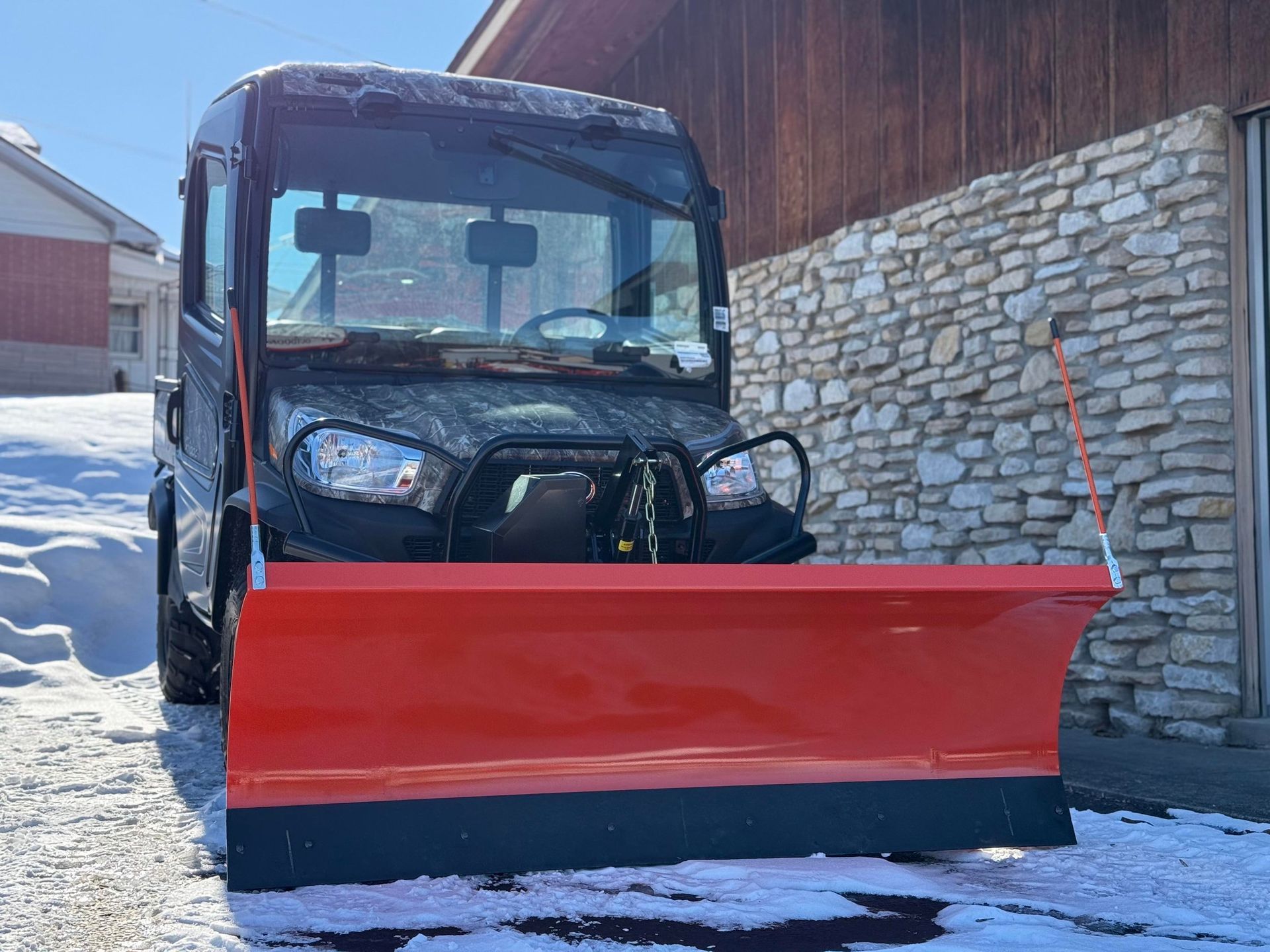 Black utility vehicle with an orange snowplow in front of a building with snow on the ground.