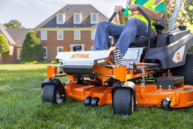 Man operating an orange STIHL riding lawnmower on a lawn, large house in background.