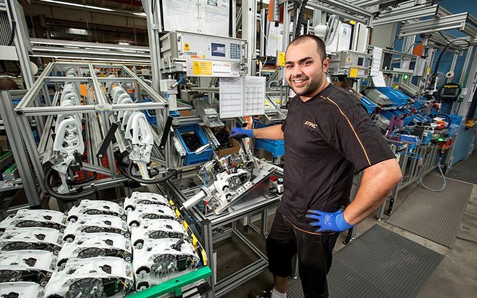 Man in factory smiles, poses with assembly line equipment; blue gloves, dark shirt.