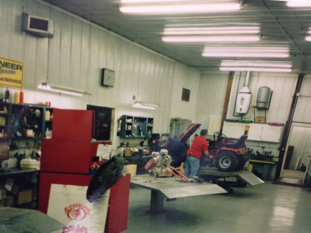 A cluttered auto repair shop with two people working on an engine and a raised vehicle.