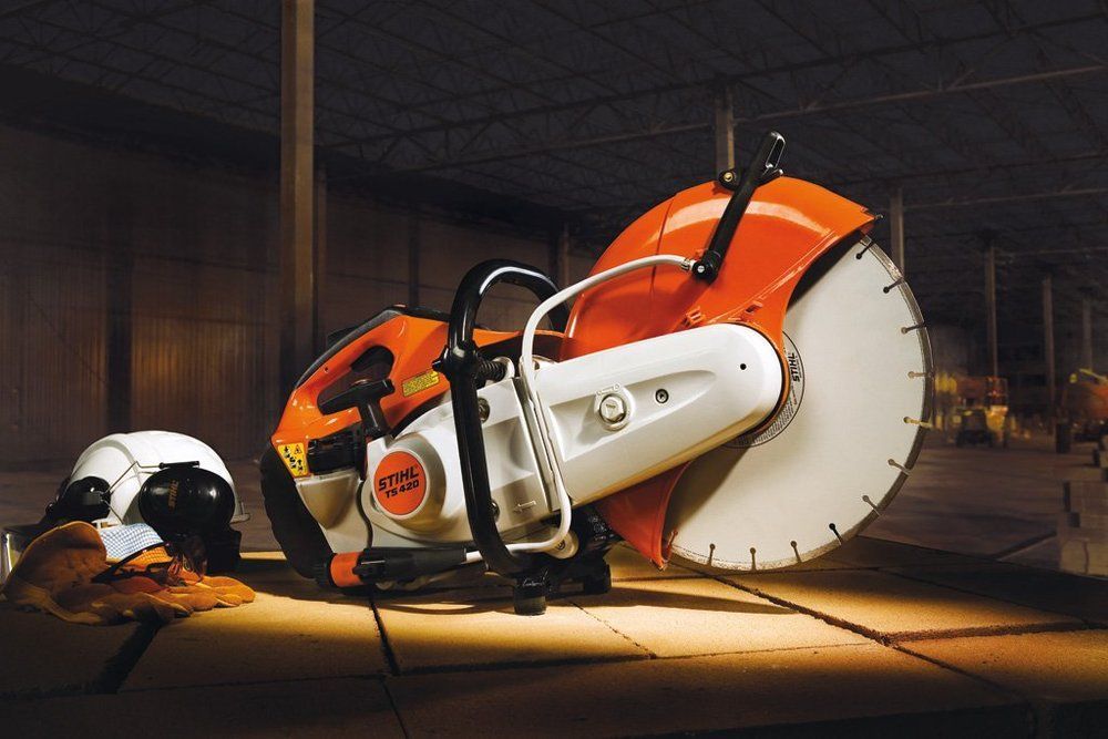 Orange and white concrete saw with a large blade, safety shield, and handle. A hardhat and gloves are beside it.