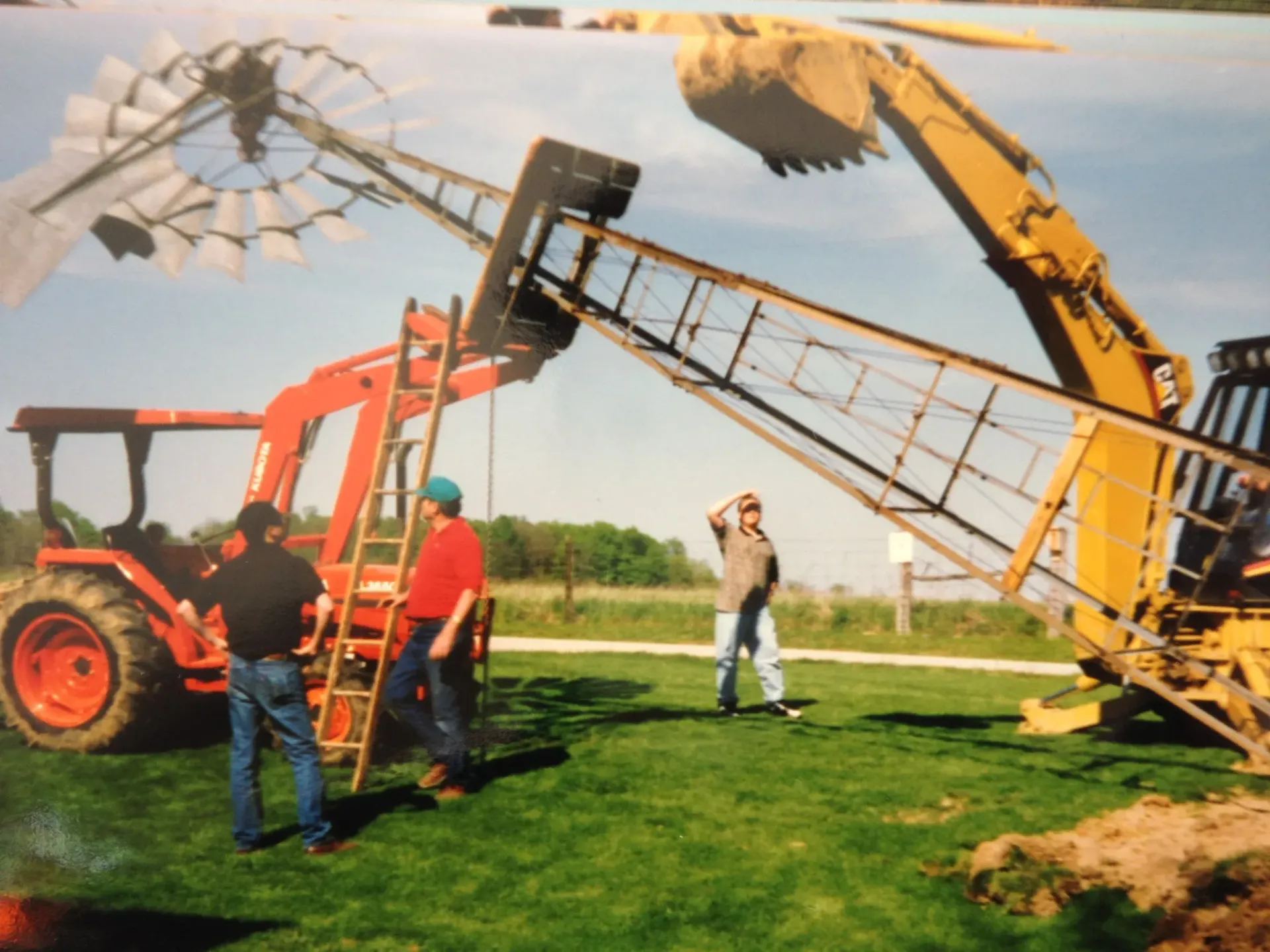 People using tractors and equipment to lower a windmill, outdoors on a grassy field.
