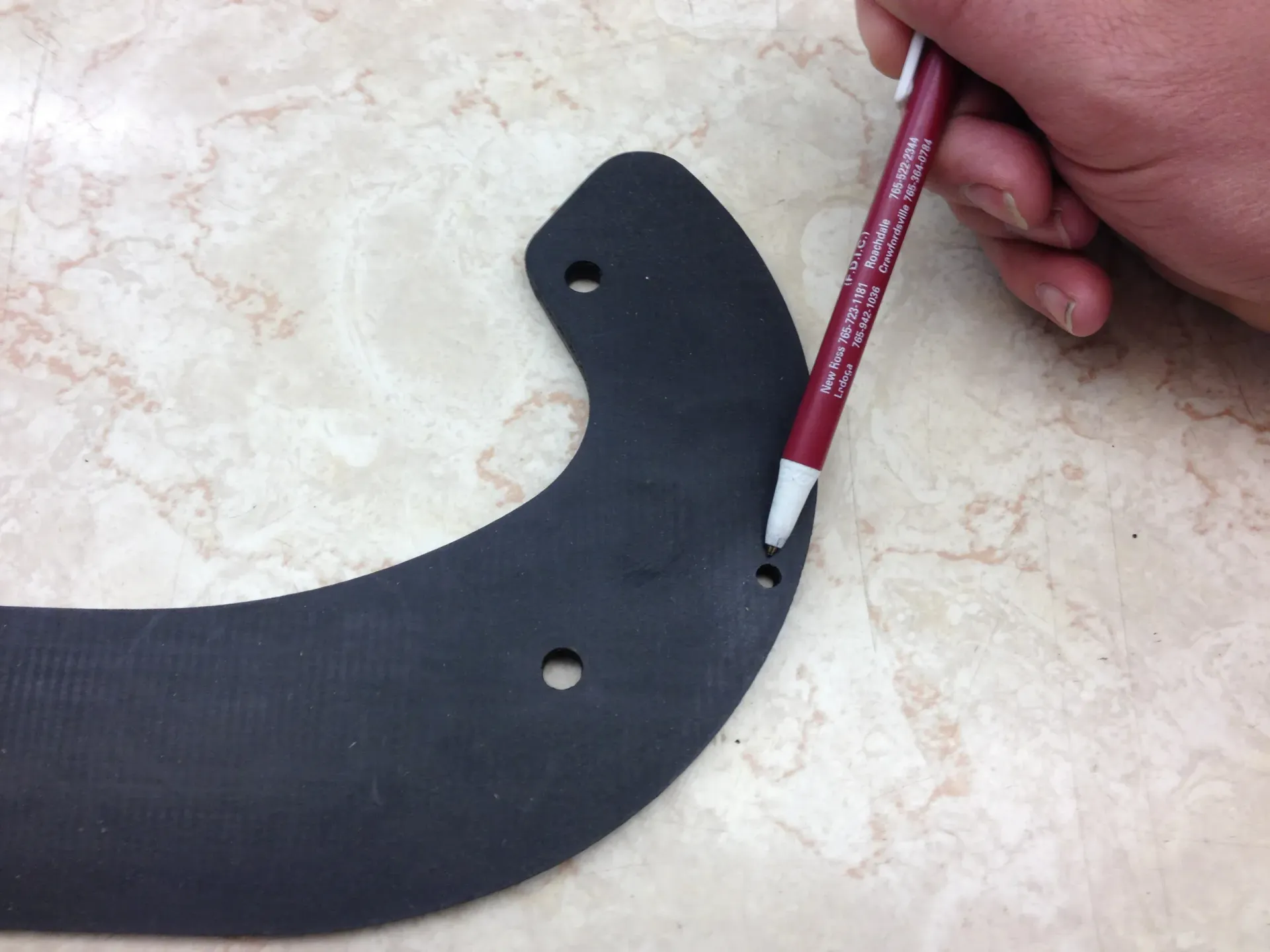 A hand using a pen to mark a hole on a black, curved plastic piece.