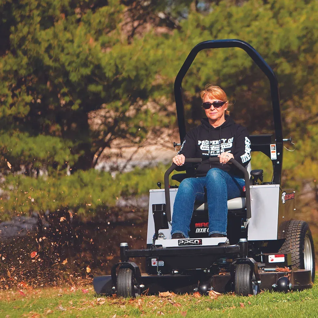 Woman on a zero-turn mower, cutting grass. She wears sunglasses and a black hoodie; leaves are blowing.