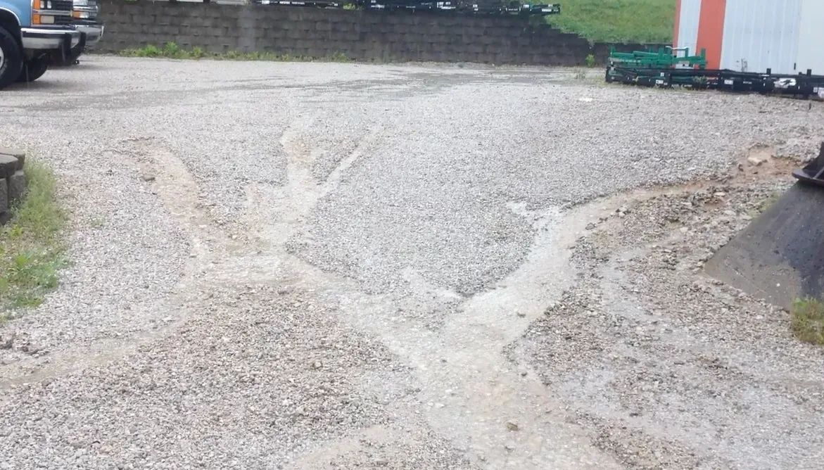 Gravel parking lot with water drainage paths, near a building and vehicles.
