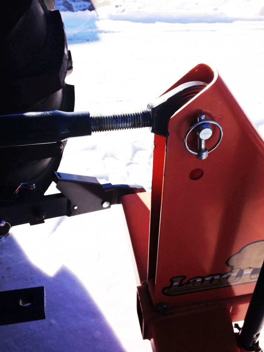 Orange snowblower attached to a tractor with a black, adjustable part and pin, set against a snow-covered background.