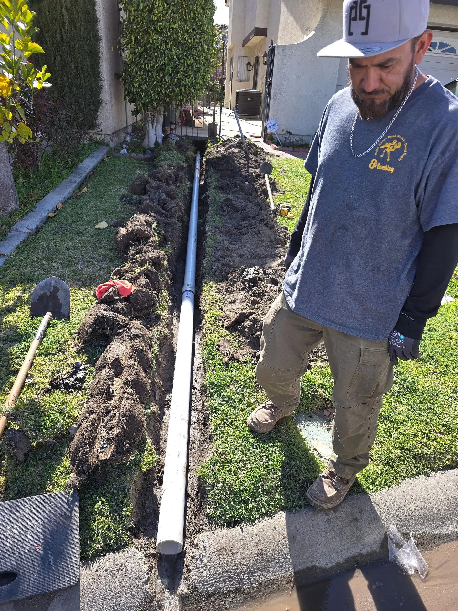 Worker standing beside a trench with a white pipe in a landscaped yard.