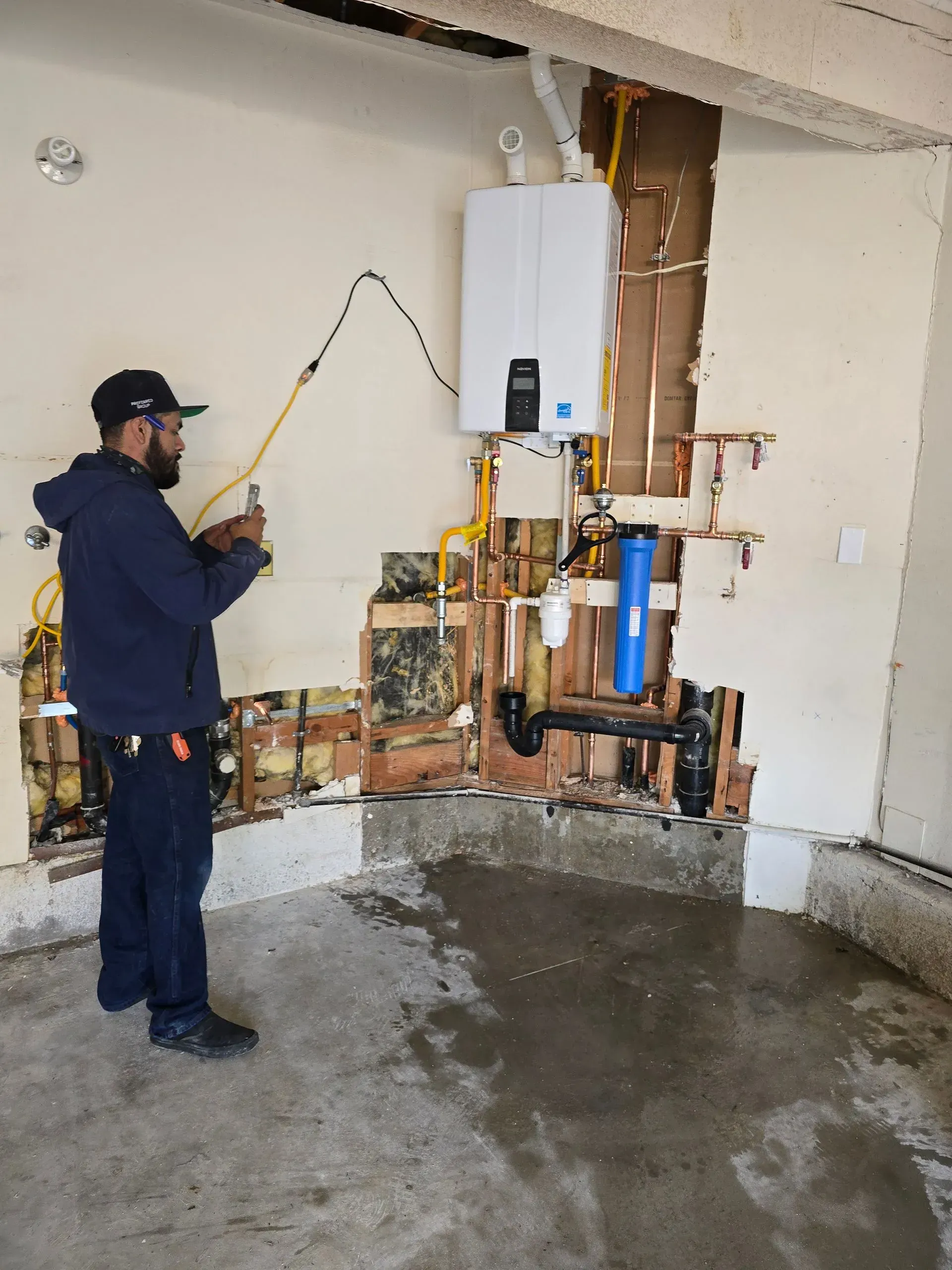 Worker inspecting wall-mounted boiler and exposed pipes in a damaged interior room
