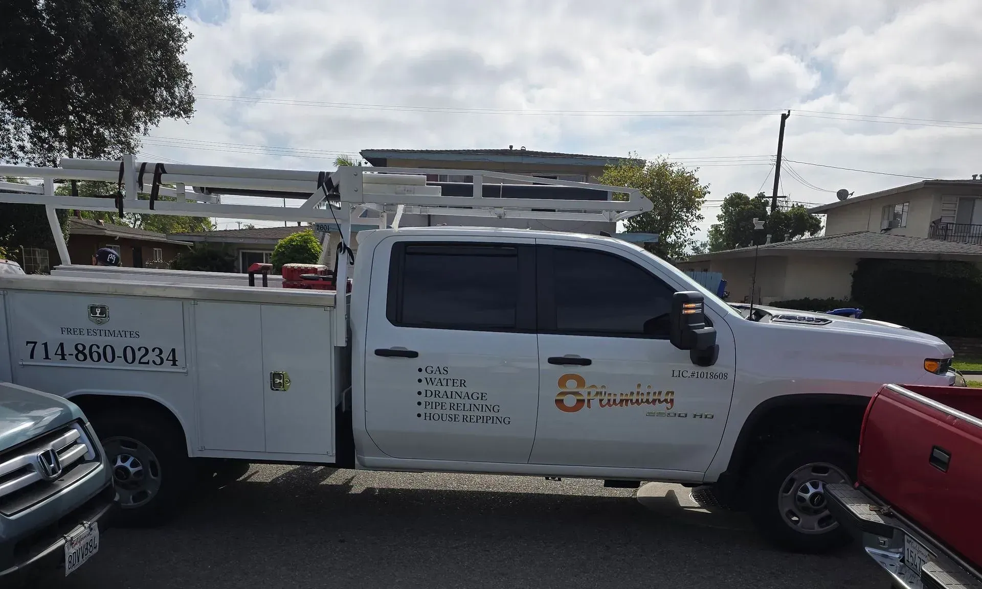 White utility truck with ladder rack parked on a street beside other cars, seen in profile