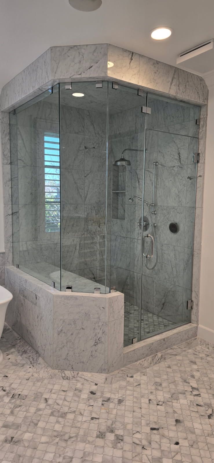 Glass-enclosed corner shower with marble tile and light stone bathroom floor
