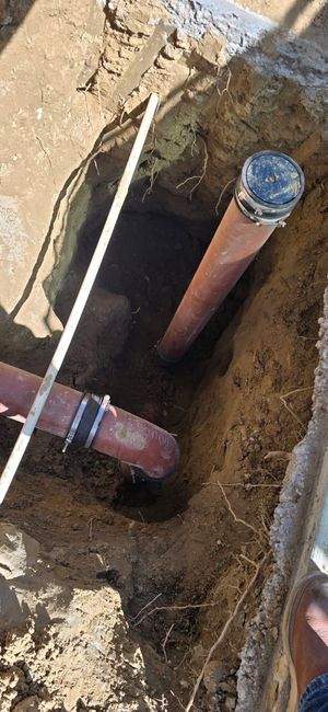 Open trench with exposed copper pipes and a valve, surrounded by dirt and construction debris