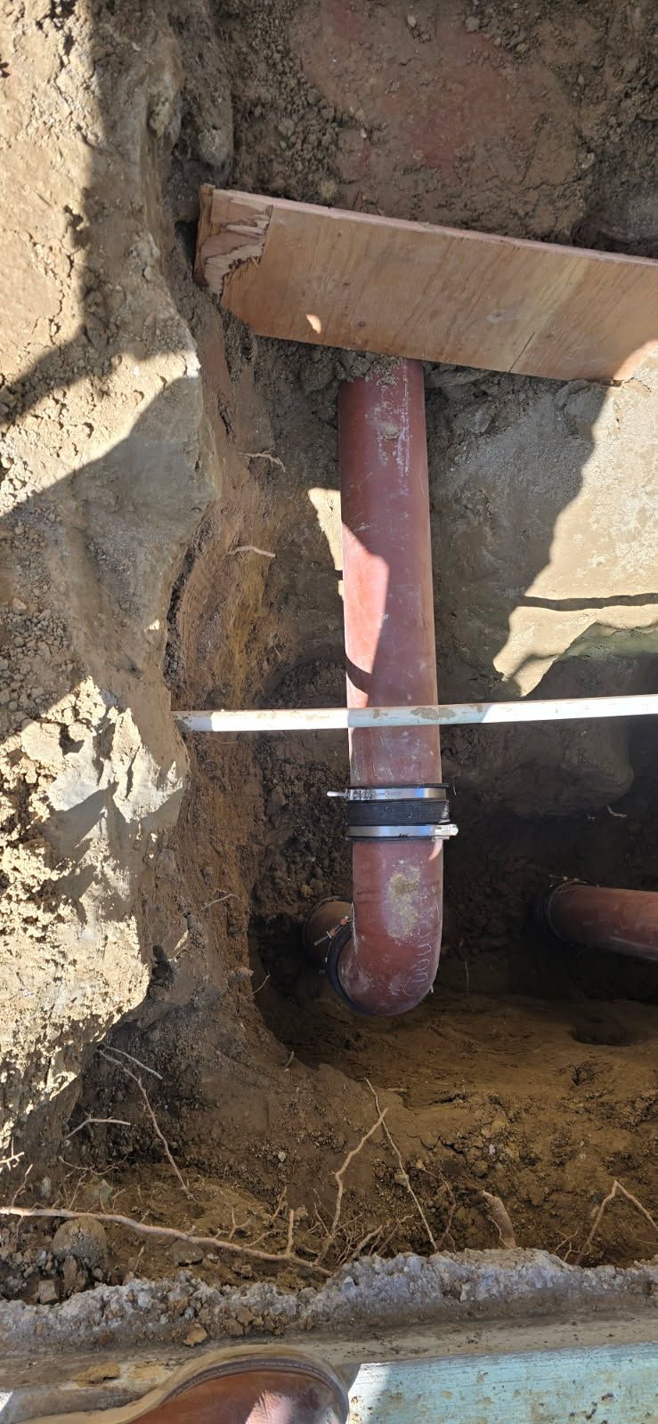 Buried red pipe with a white crossbar, exposed in a dirt trench next to a wooden board.