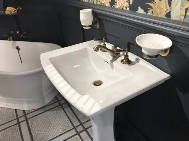 Bathroom sink with white pedestal basin, faucet, toilet, and bathtub in a dark-walled room with floral wallpaper.