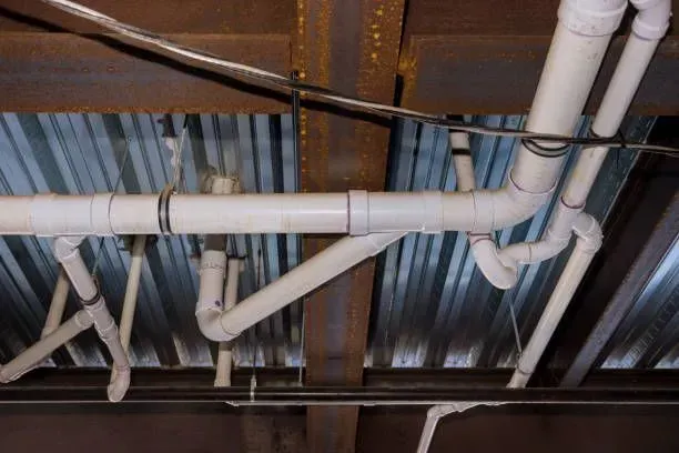 Exposed white PVC plumbing pipes under a corrugated metal ceiling with wooden joists.