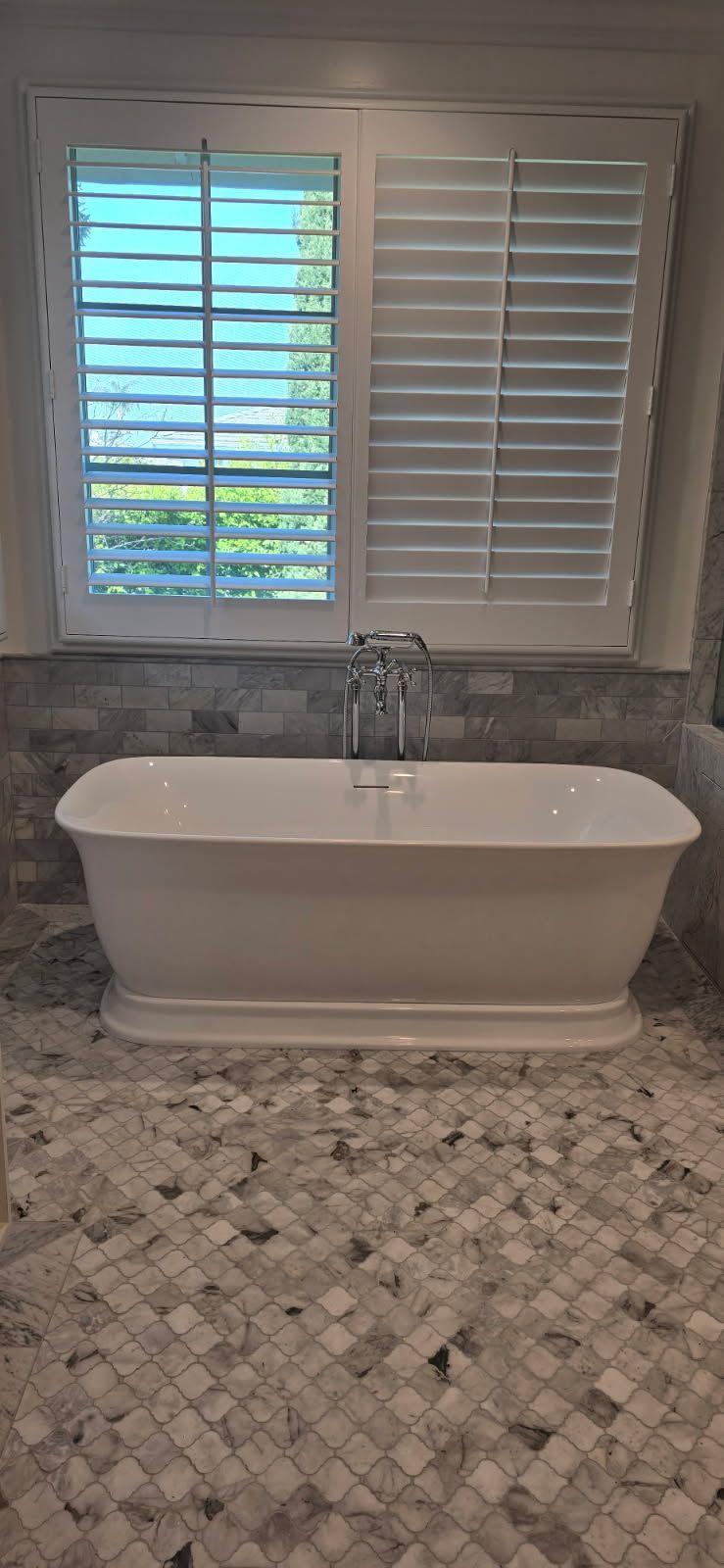 White freestanding bathtub beneath a window with shutters in a tiled bathroom