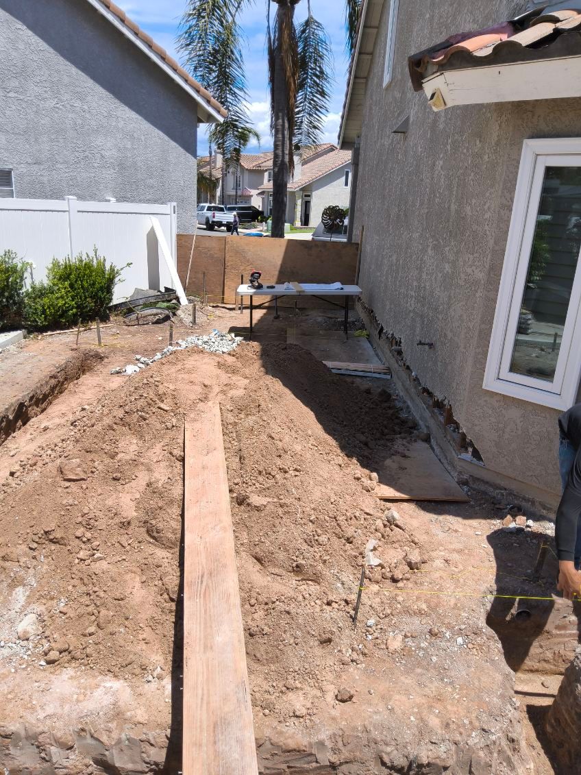 Narrow dirt trench along a house exterior with tools and construction materials in a backyard.