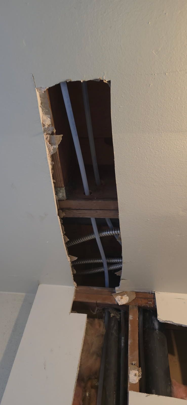 Damaged ceiling with a large hole exposing beams and wiring above a white wall.