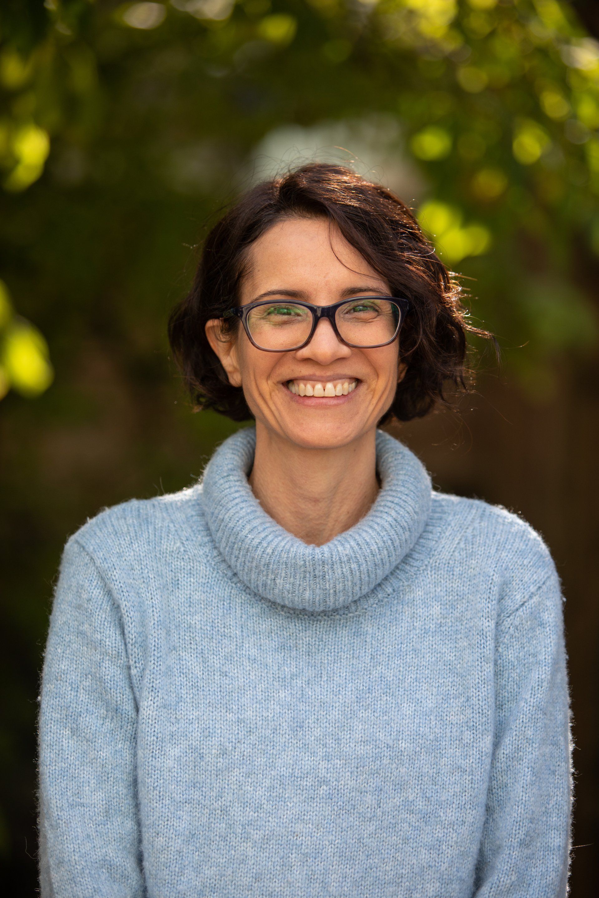 Smiling person wearing glasses and a light blue sweater, standing outdoors against a blurred green background