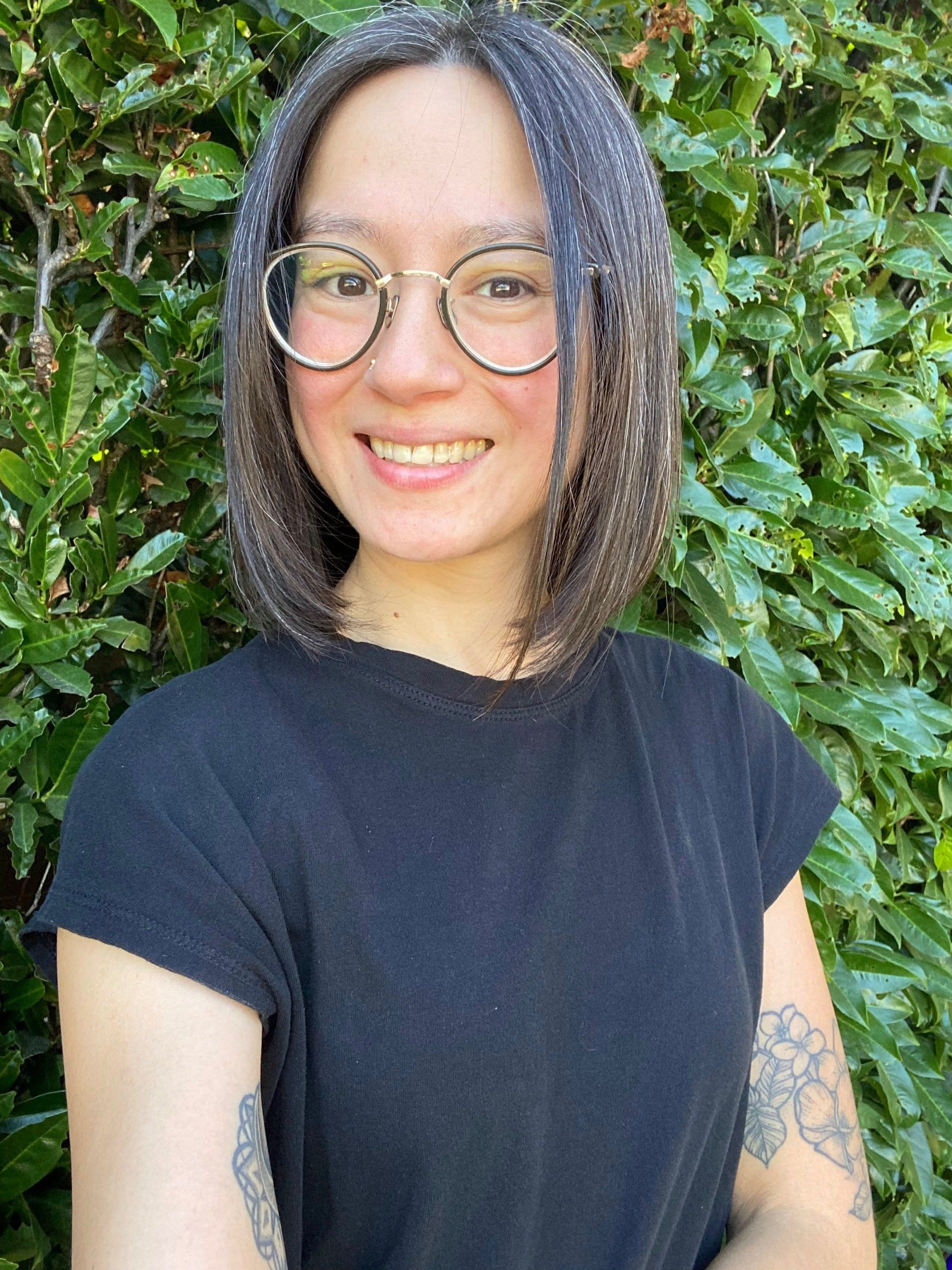 Smiling person with glasses and a black shirt standing in front of green foliage