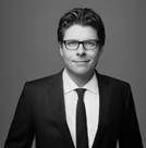 Man in suit and glasses looking to the side with a slight smirk.