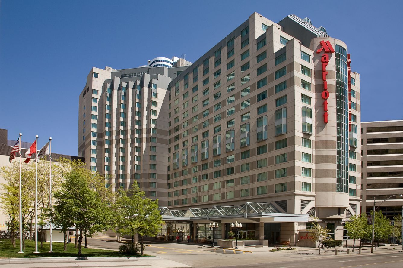 A multi-story Marriott hotel building with a glass entrance canopy, surrounded by green trees under a clear blue sky.