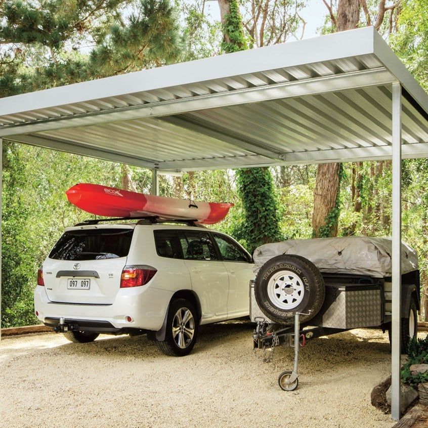 A white SUV with a red kayak on its roof is parked under a metal carport next to a small utility trailer in a wooded area.