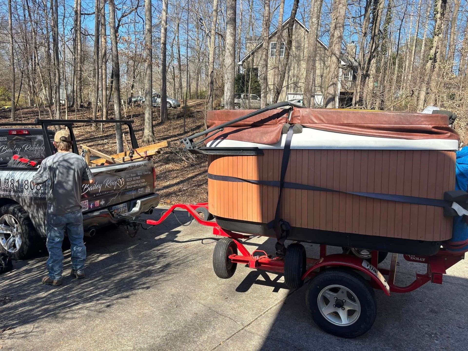 A man loads a hot tub on a trailer, attached to a pickup truck, in a wooded area.