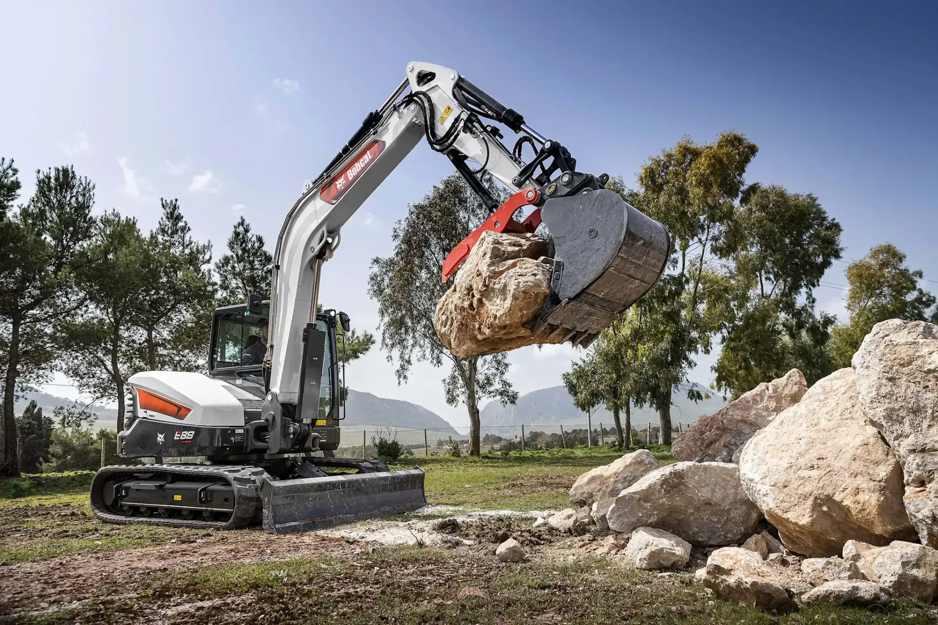 Escavatore Bobcat con benna a pinza che solleva una grande roccia.