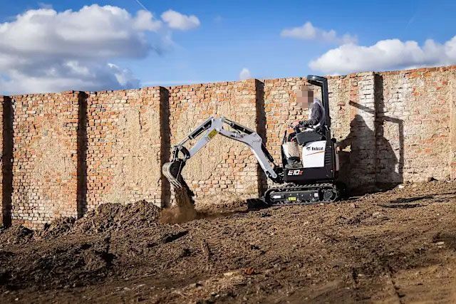 Miniescavatore che scava terra vicino a un muro di mattoni.