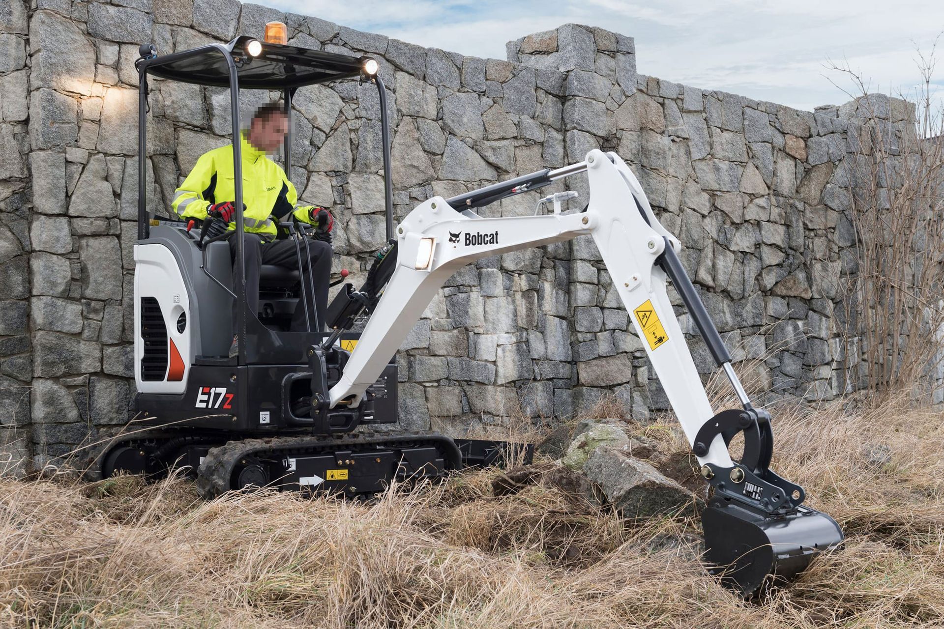 Persona che utilizza un miniescavatore Bobcat mentre scava vicino a un muro di pietra.