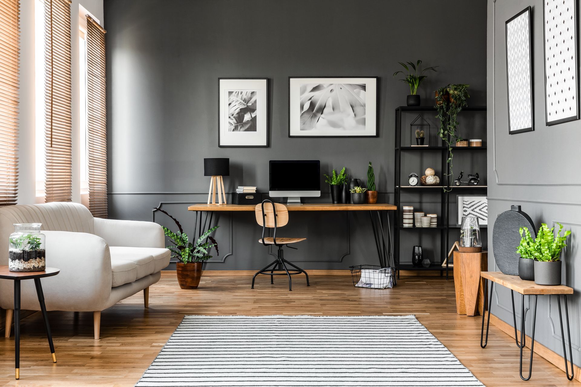 A stylish home office with gray walls, wood floors, a desk with a computer, a sofa, and potted plants.