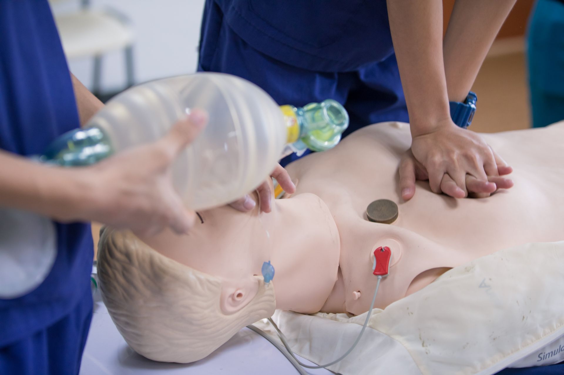 People practicing CPR on a training mannequin; one uses a bag valve mask, the other does chest compressions.