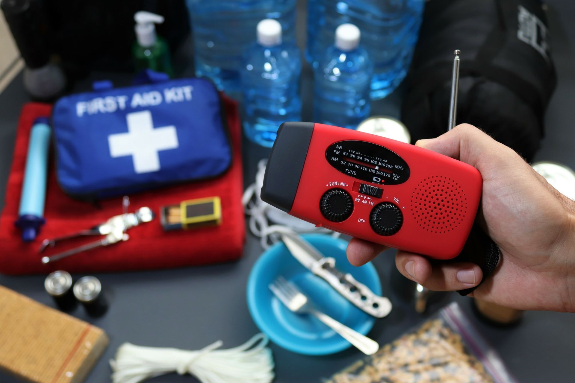 Hand holding a red emergency radio, surrounded by a first aid kit, water bottles, and other survival supplies.