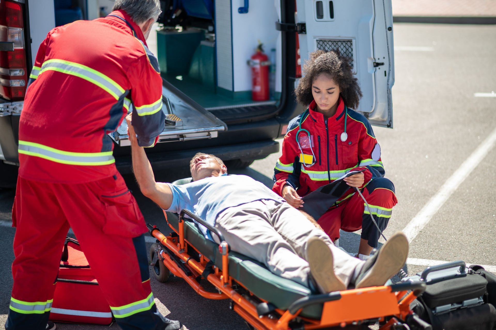 Paramedics loading a person on a stretcher into an ambulance. One kneels, checking vitals; other assists.