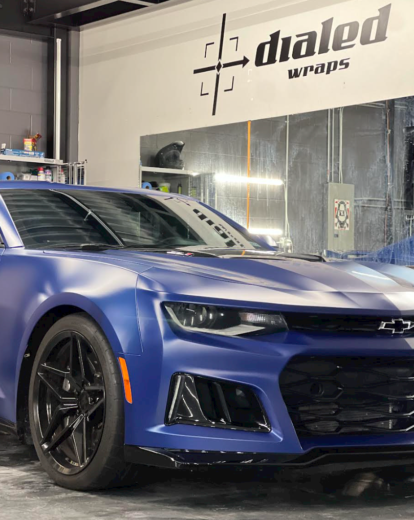 A blue car is parked in front of a wall that says dialed wraps