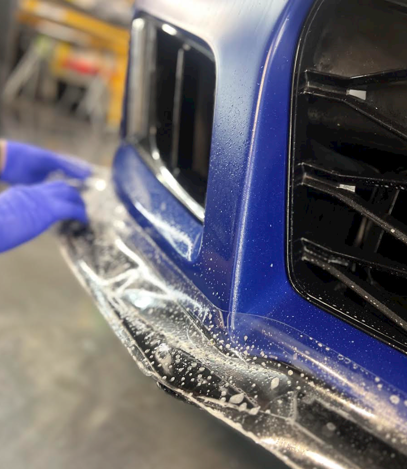 A person is applying plastic wrap to the front bumper of a blue car.