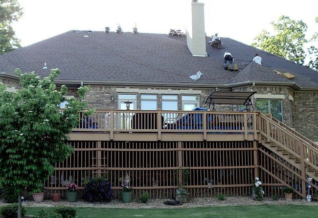 Roofers working on a dark shingled roof of a brick house with a wooden deck and green backyard.