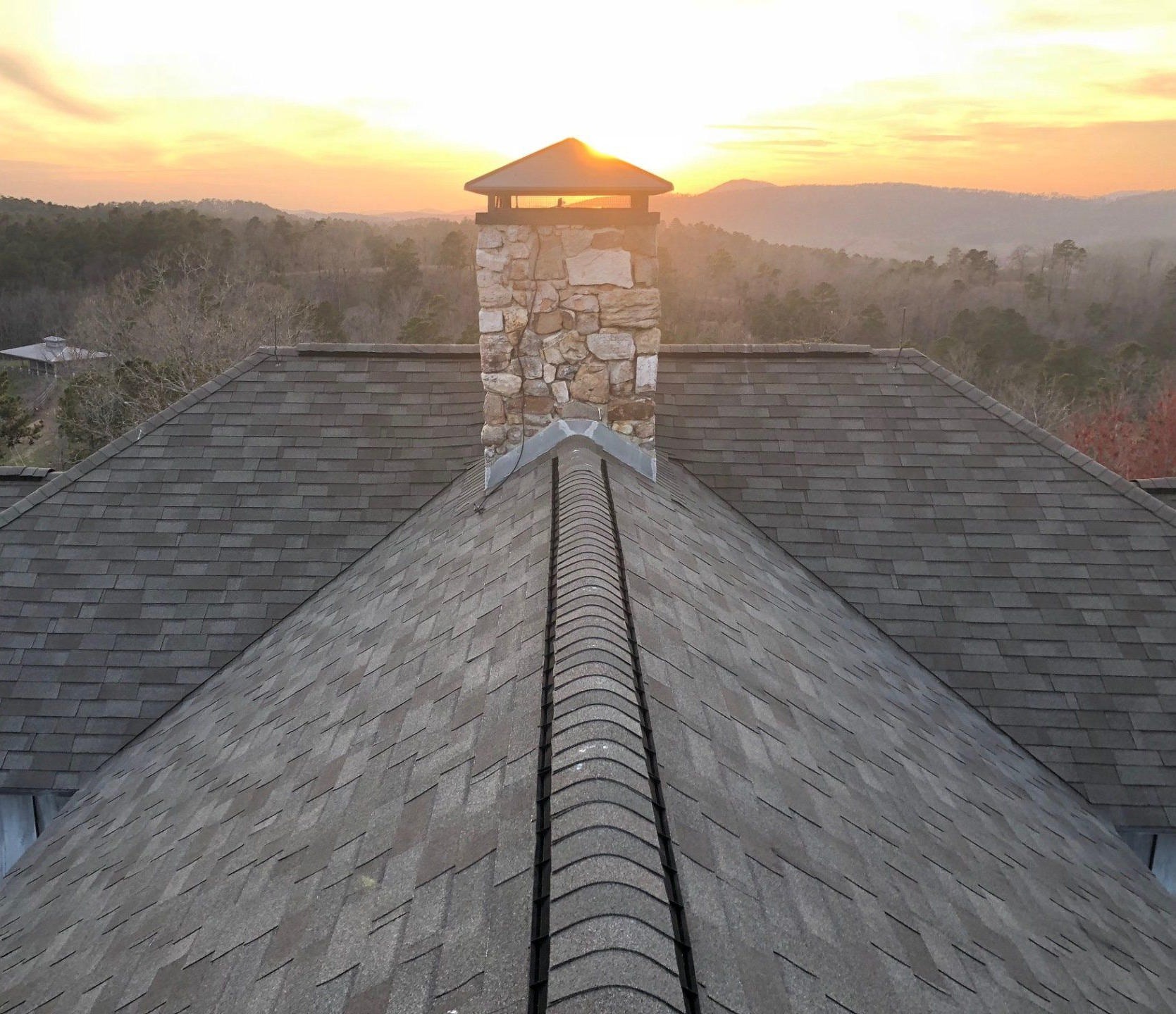 Rooftop view of a chimney, sunset in the background, gray shingles, stone chimney, and mountain view.