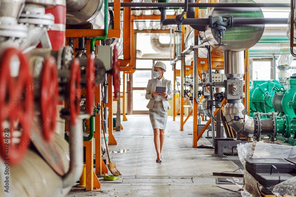 Woman in a hard hat and suit walks through an industrial facility, inspecting equipment and holding a tablet.