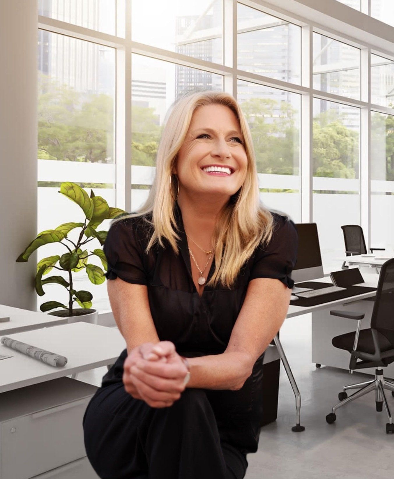 Blonde woman smiles, seated on a desk in a bright office; arms crossed.