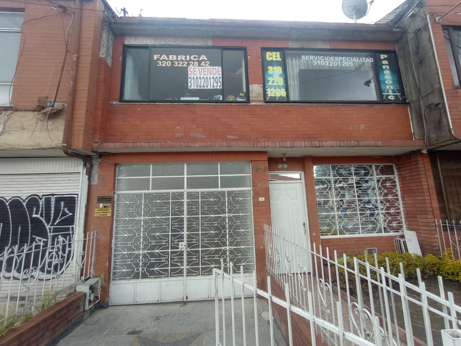 Fachada de ladrillo rojo con ventanas enrejadas, grafitis y una valla metálica blanca delante.