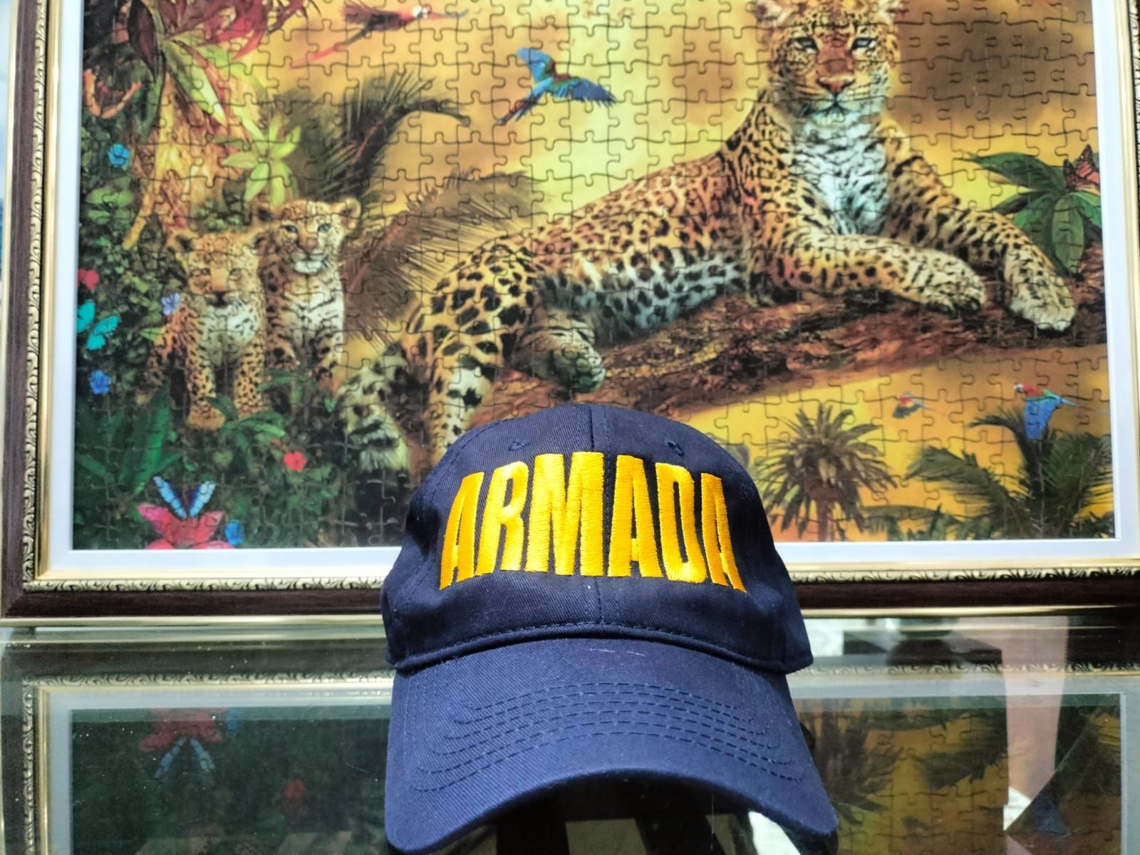 Gorra azul con texto amarillo “ARMANI” frente a una lámina decorativa con estampado de leopardo en la selva.