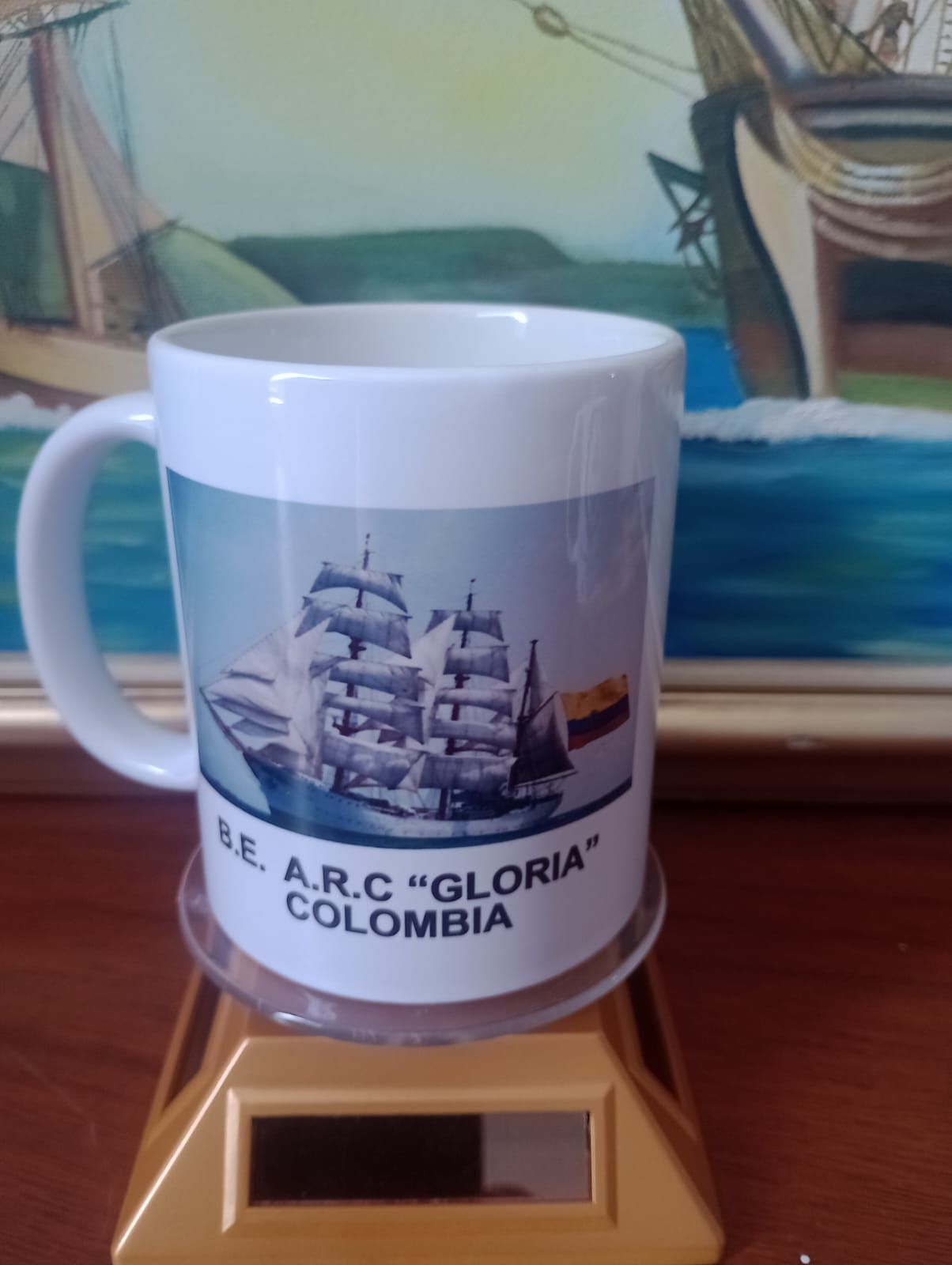 Taza blanca con la ilustración de un barco y el texto “ARC GLORIA COLOMBIA”, sobre un pequeño soporte de madera.