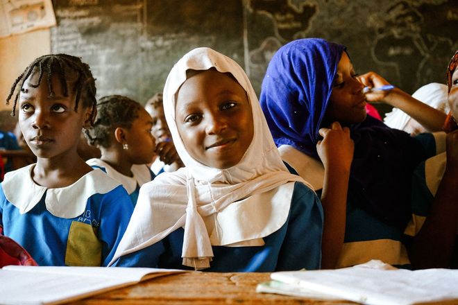Schoolchildren seated in a classroom, with one girl in a white headscarf smiling at the camera.