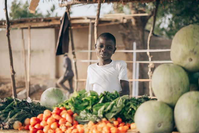 Child behind a market stall with fresh tomatoes, greens, and melons outdoors