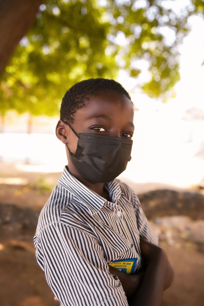 Child wearing a black face mask and striped shirt standing outdoors in front of a tree