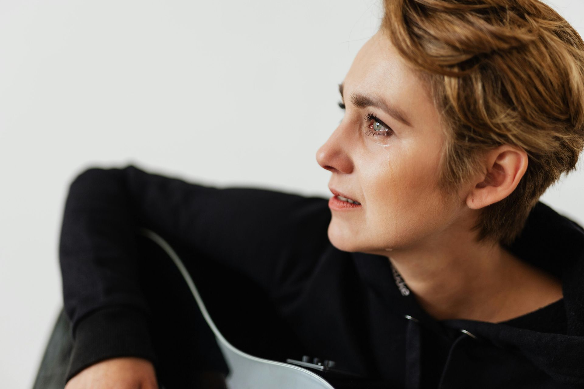 Person in a black top holding a guitar, looking to the side against a white background